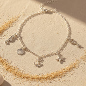Custom charm anklet displayed on sand with circular arrangement, sterling silver anklet with multiple dangling charms, personalized ankle bracelet handmade for women, minimalist charm jewelry inspired by beach and summer style, adjustable silver anklet designed for comfort and layering, lightweight charm anklet perfect for vacation jewelry, seaside wear, and meaningful personalized gifts