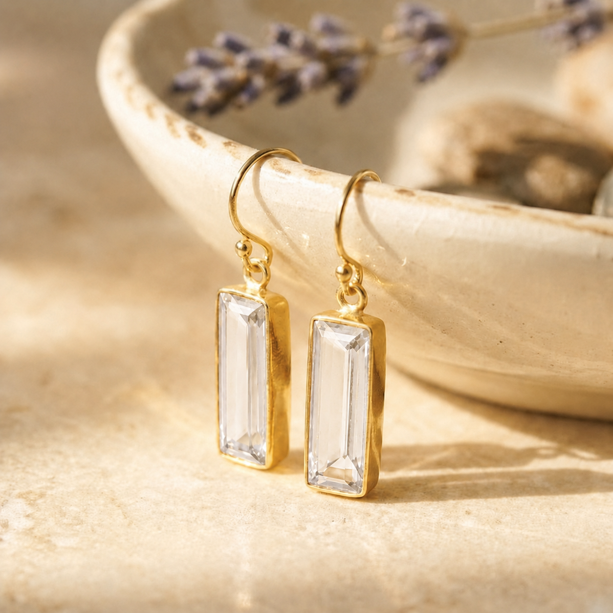 Clear quartz bar drop earrings in yellow gold vermeil displayed on a ceramic bowl in warm natural sunlight.