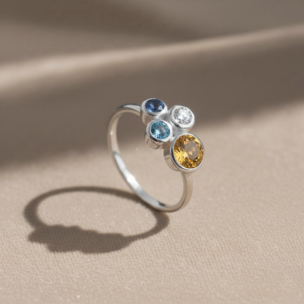 three stone birthstone ring resting on champagne satin fabric with warm light highlighting the gold vermeil finish and vibrant gemstones, personalized mom ring with three birthstones representing each child, elegant cluster style ideal for everyday wear or special occasions, thoughtful Mother’s Day gift, birthday gift or family jewelry heirloom crafted in sterling silver or gold vermeil with customizable stones