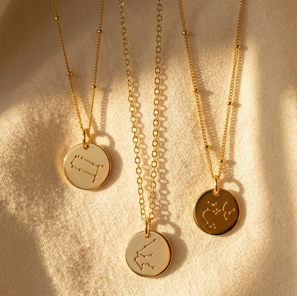 Zodiac constellation necklaces in sterling silver, yellow gold, and rose gold finishes on a neutral fabric background.