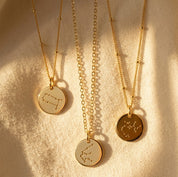 Zodiac constellation necklaces in sterling silver, yellow gold, and rose gold finishes on a neutral fabric background.
