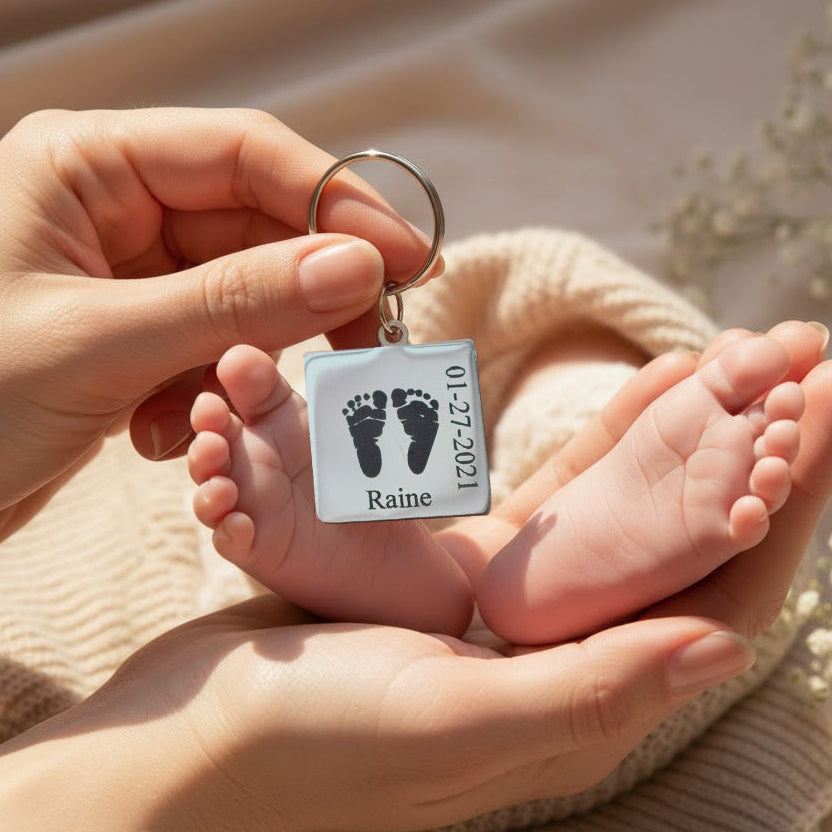 Custom engraved baby footprint keychain resting against a newborn’s tiny feet, held in a parent’s hands on a soft blanket, showing the square metal tag with realistic footprints, baby name and birth date. Personalized new baby gift or memorial keepsake for mom, dad or grandparents, perfect for first Father’s Day, Mother’s Day, baby shower or hospital birth announcement.