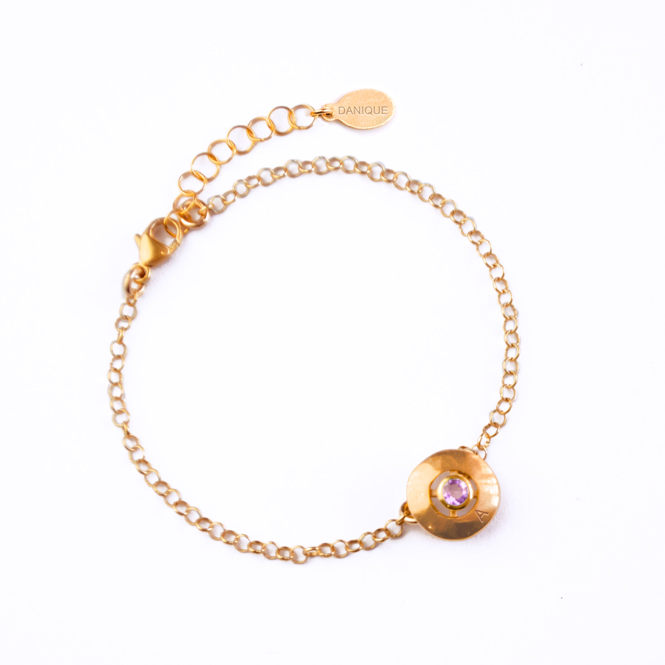 A rose gold disc bracelet with the letter "A" engraved at the bottom with a purple amethyst in the center rests against a white background. This is perfect for February birthday babies, Moms who want to keep their children's birthstones close to her, and anyone looking to add a meaningful bit of sparkle to their outfits. 