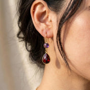 Woman wearing purple and red gemstone double drop earrings with gold ear wires.