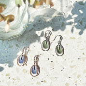 Two pairs of open oval drop earrings with hook backings and a textured oval disc at the center plated in either rhodium with green enamel or rose gold with blue enamel rest on a textured white stone background next to a plant and a glass.