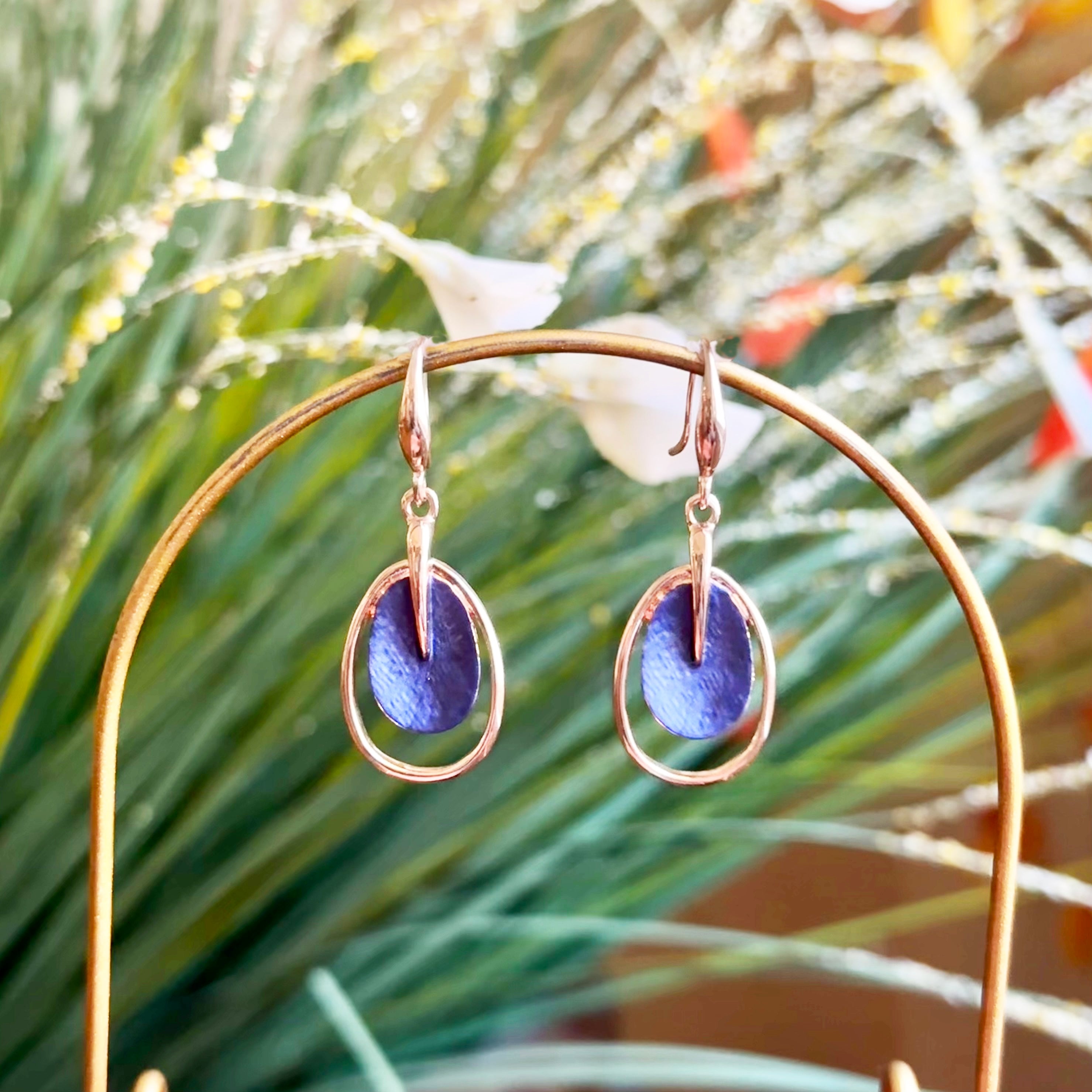 A pair of rose gold plated open oval drop earrings with hook backings and a textured oval disc at the center painted in blue enamel rest on rounded copper wire with plants behind them