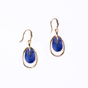 A pair of rose gold plated open oval drop earrings with hook backings and a textured oval disc at the center painted in blue enamel rest on rounded copper wire with plants behind them