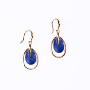 A pair of rose gold plated open oval drop earrings with hook backings and a textured oval disc at the center painted in blue enamel rest against a white background.