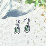 A pair of rhodium plated open oval drop earrings with hook backings and a textured oval disc at the center painted in green enamel rest on a white and gray color block stone coaster.