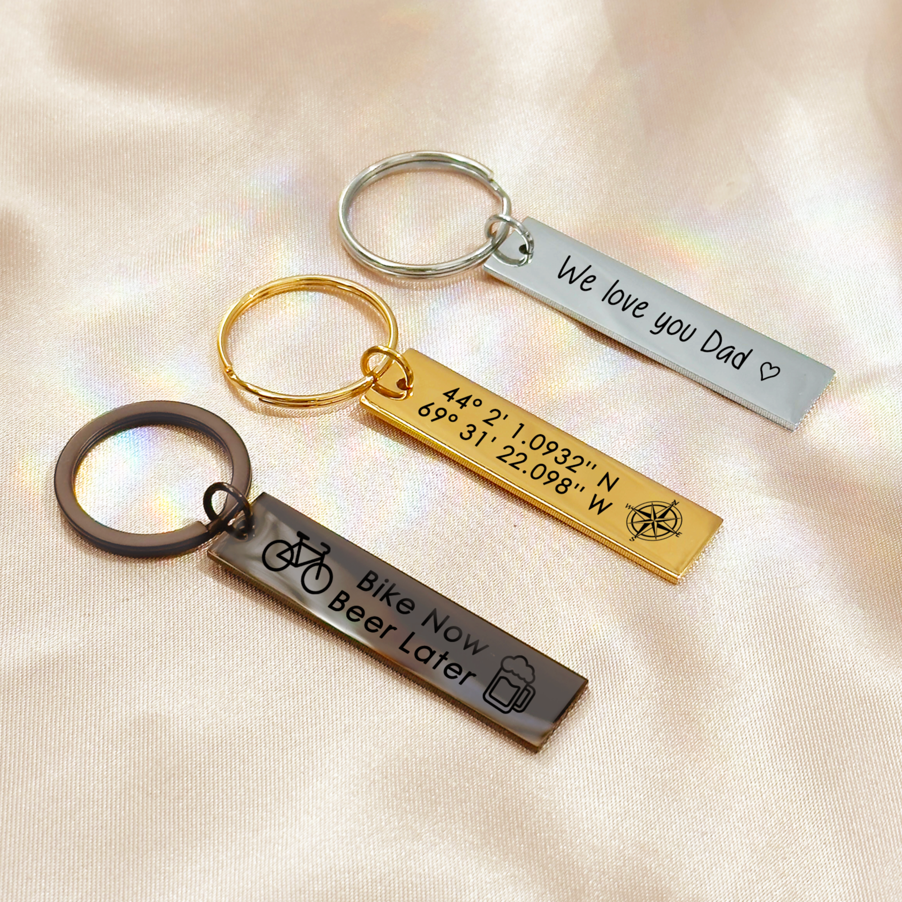 Personalized engraved bar keychains in silver, gold, and black finishes displayed on satin background, featuring custom text and symbols. Designs include “We love you Dad” with heart outline, custom GPS coordinates with compass engraving, and “Bike Now Beer Later” with engraved bike and beer mug icons. Customizable stainless steel bar keychains make meaningful gifts for Father’s Day, birthdays, anniversaries, or everyday keepsakes for men, dads, cyclists, and loved ones.