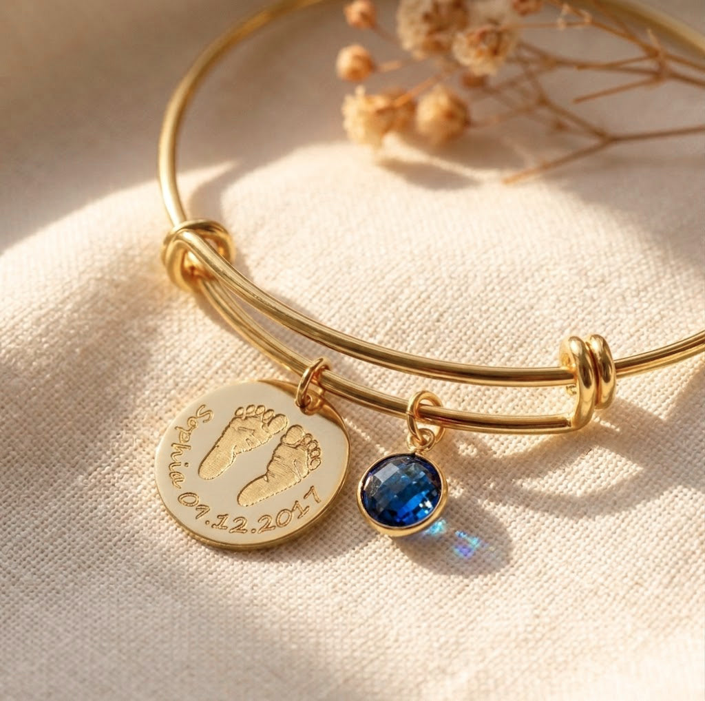 Close-up of custom baby footprint bangle with engraved name, date, and blue birthstone charm on gold adjustable bracelet.