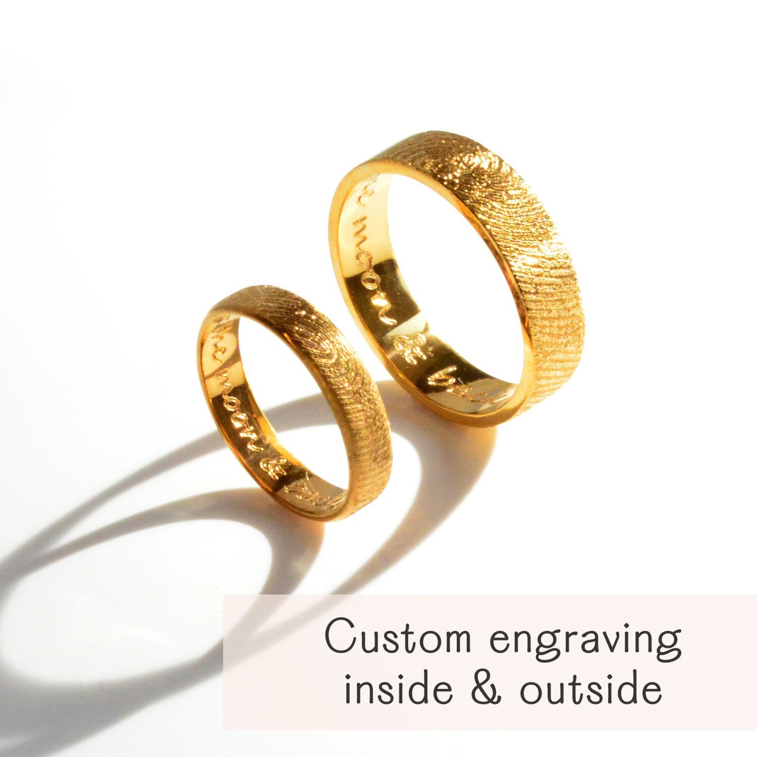 Two gold fingerprint rings with textured outside band and shiny inner surface engraved with custom handwritten message, shown on white background with shadows and text “custom engraving inside & outside,” personalized couples wedding bands or promise rings, unique keepsake jewelry gift.