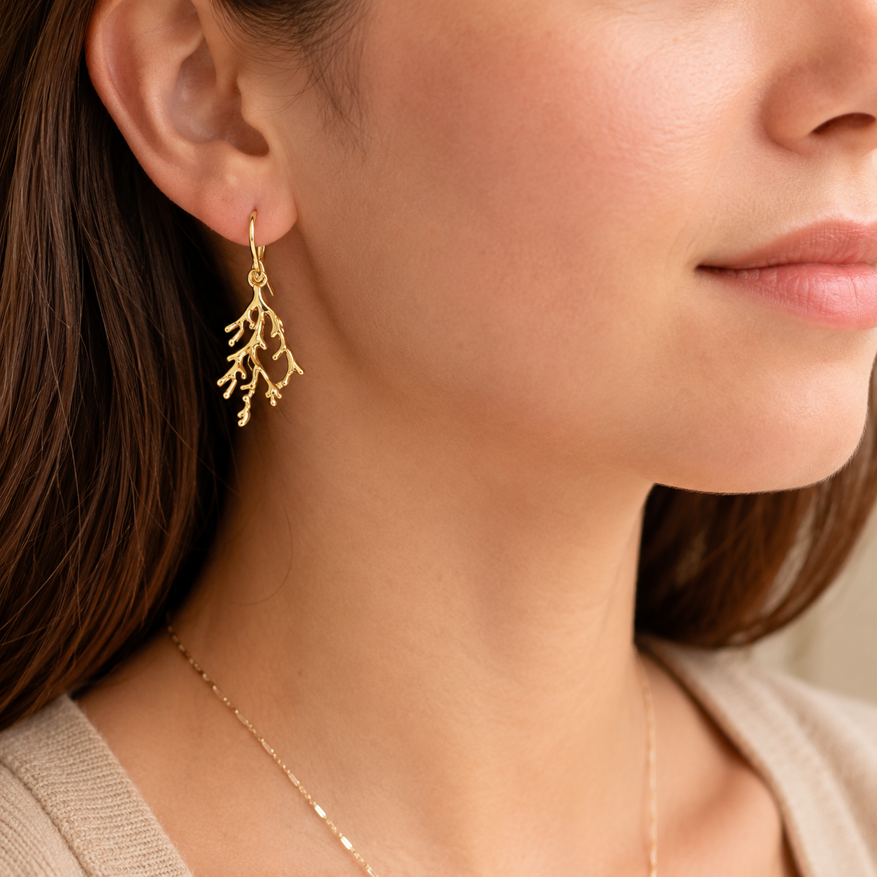A close-up of a woman wearing a delicate gold drop earring shaped like branching coral or seaweed, paired with a thin gold necklace and a beige top.