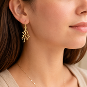 A close-up of a woman wearing a delicate gold drop earring shaped like branching coral or seaweed, paired with a thin gold necklace and a beige top.