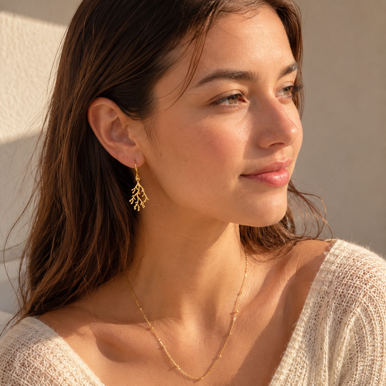 A woman wearing delicate gold leaf dangle earrings and a fine gold chain necklace, shown in a warm, close-up lifestyle portrait.