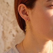 A close-up of a woman's ear wearing a delicate gold dangle earring with a leaf or branch-like organic design, set against a softly blurred warm beige background.
