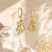 A pair of gold drop earrings with a coral or branch-like organic structure, hanging against a soft beige fabric backdrop with warm sunlight and dried flowers.