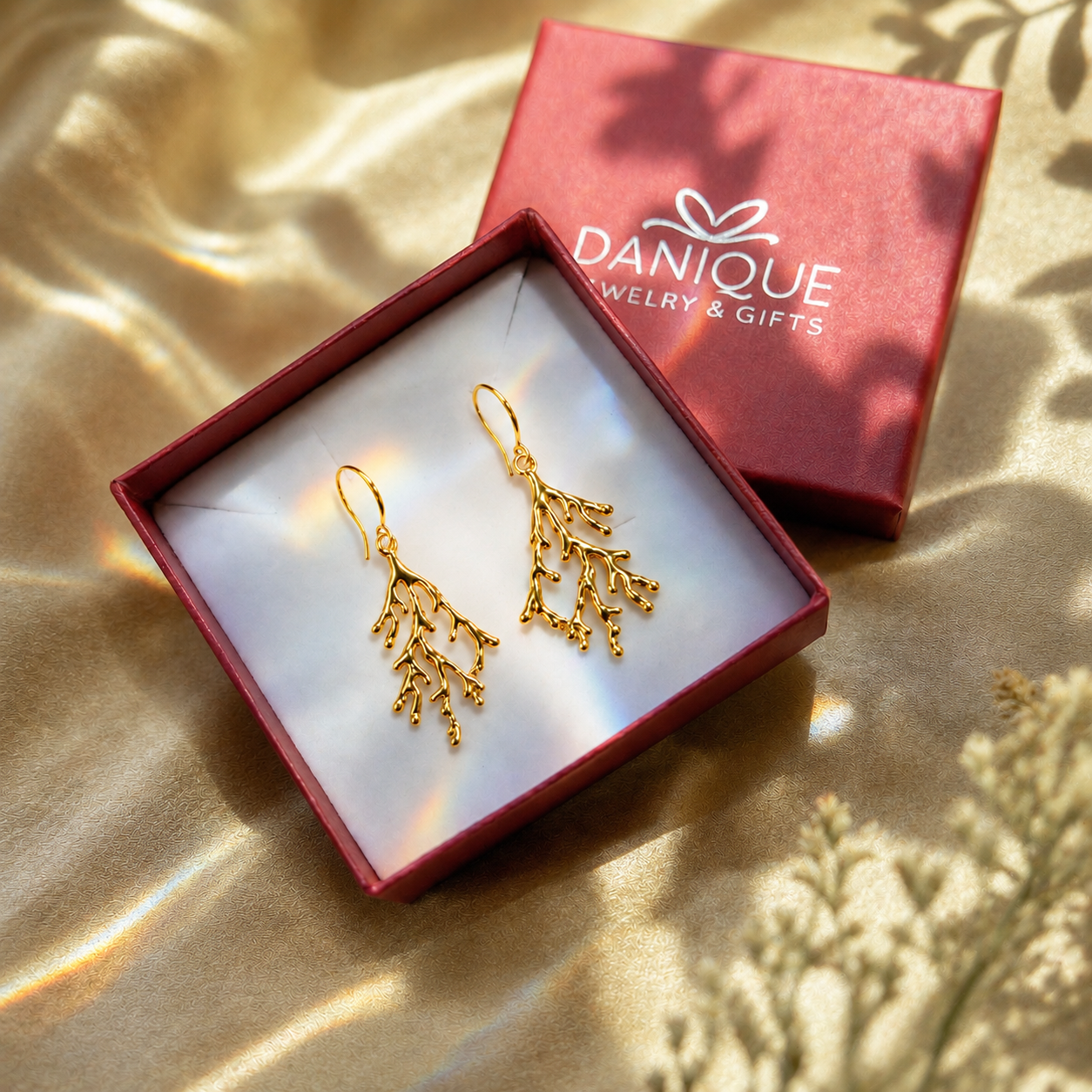 A pair of gold branch-shaped dangle earrings displayed in an open red jewelry box, with the matching red Danique Jewelry & Gifts box placed behind it on a shiny gold fabric background.