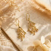 A pair of gold branch-shaped dangle earrings displayed on a textured beige stone surface with dried flowers and soft natural shadows.