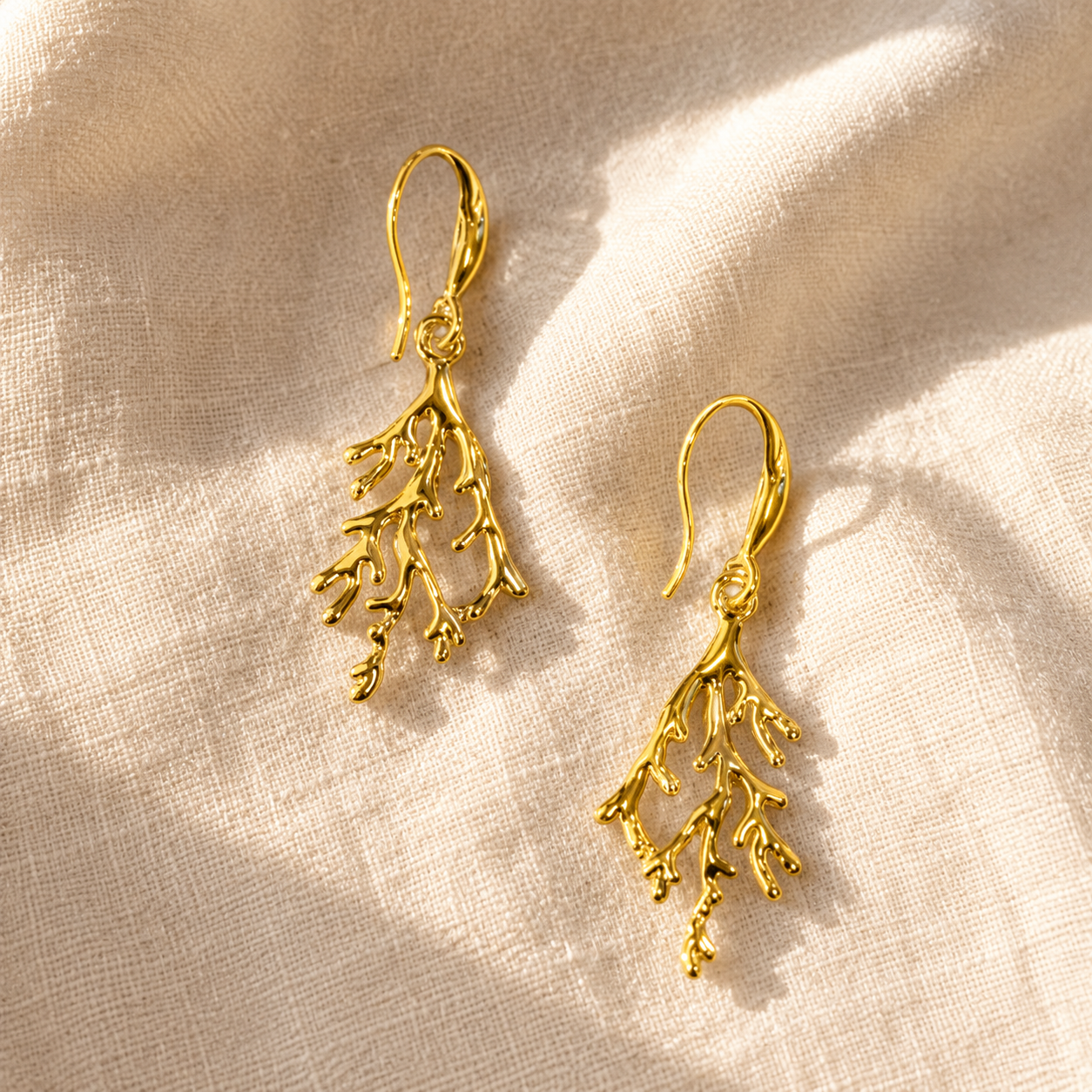 A pair of gold drop earrings with a delicate branch or coral-like leaf design, displayed on a textured beige fabric background with soft shadows.