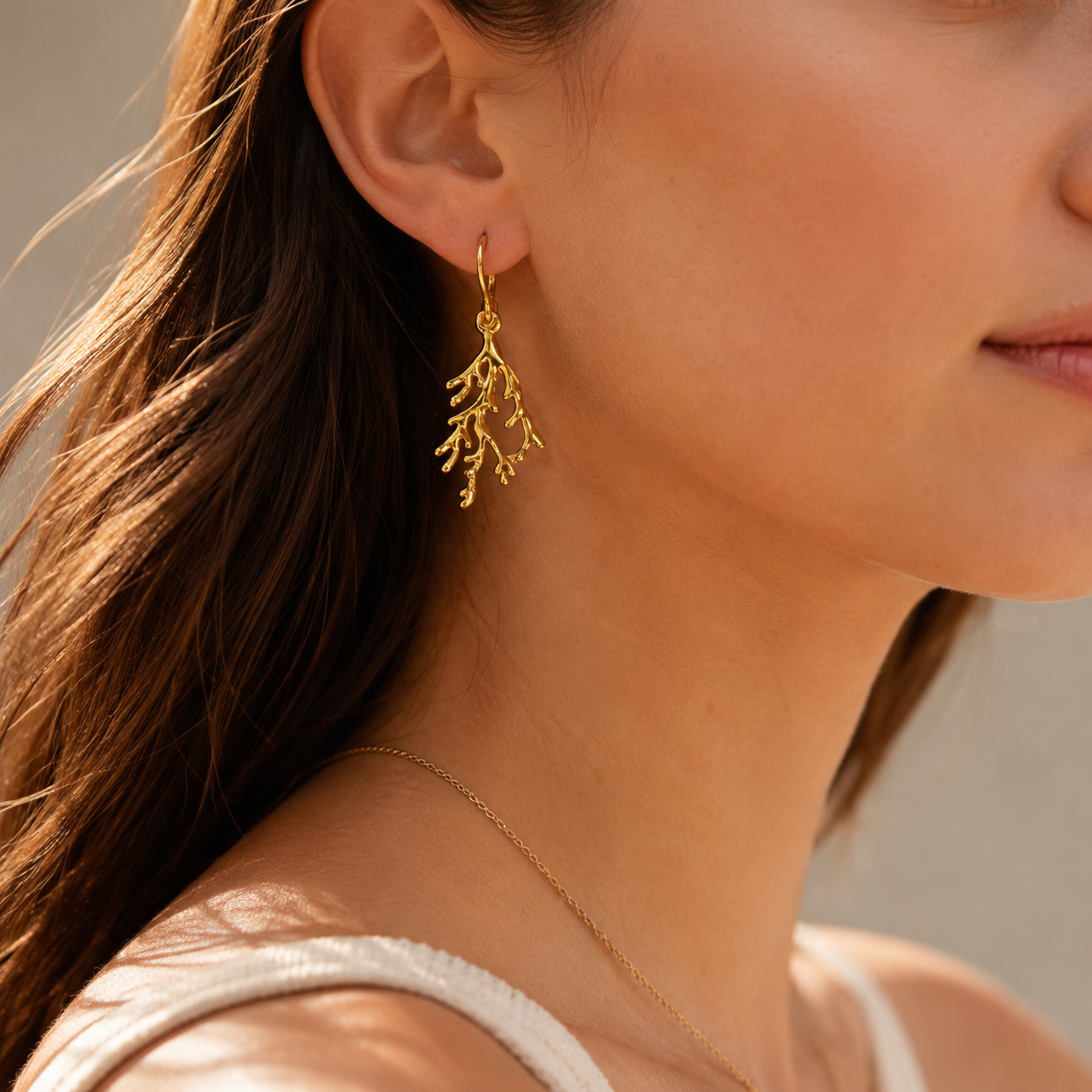 A close-up of a woman wearing a delicate gold dangle earring with a coral-like branch design, paired with a thin gold chain necklace and a white top.