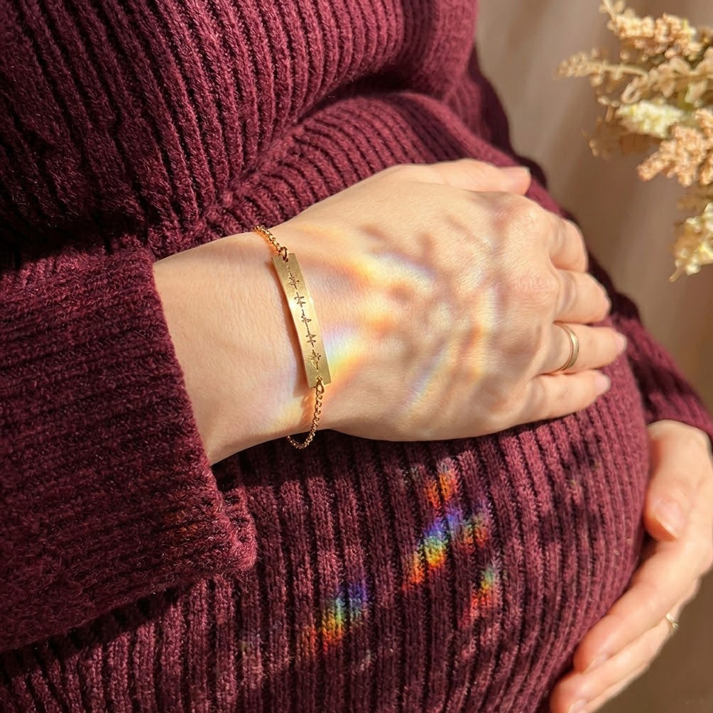 Custom gold bar bracelet with engraved heartbeat design worn by an expecting mother resting her hand on her pregnant belly, meaningful pregnancy jewelry symbolizing baby’s heartbeat; elegant personalized bracelet for maternity photoshoot, baby shower gift, mama-to-be present, sentimental keepsake jewelry for new mom, delicate gold chain accessory with minimalist engraved medical heartbeat motif.