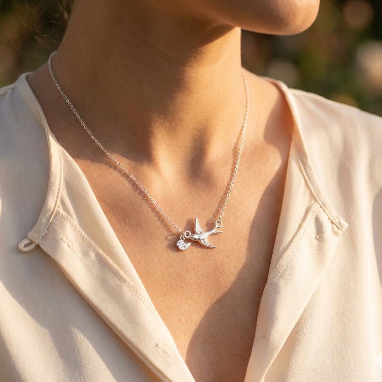 Woman wearing a silver mama bird necklace with tiny baby bird charms on a delicate chain, photographed in warm sunlight. Minimalist family jewelry symbolizing motherhood, children, and love.