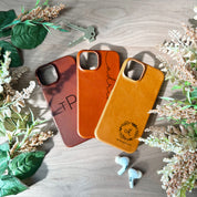 Three engraved leather phone cases rest on a wood background. A pair of airpods are below the right most iPhone case. There are flowers on the edges and a set of keys above the left most iPhone cases. These iPhone 15 and other sized cases are perfect for adding a touch of personalization to your daily accessories. Engraved leather phone cases, phone case for iPhone 15, custom engraved name monogram phone accessory.