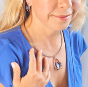 Adjustable Necklace with a brown steel wire chain and an open Pear shape rose gold pendant with a floating textured disc center painted in blue enamel with matching stud earrings worn by a model in a blue blouse