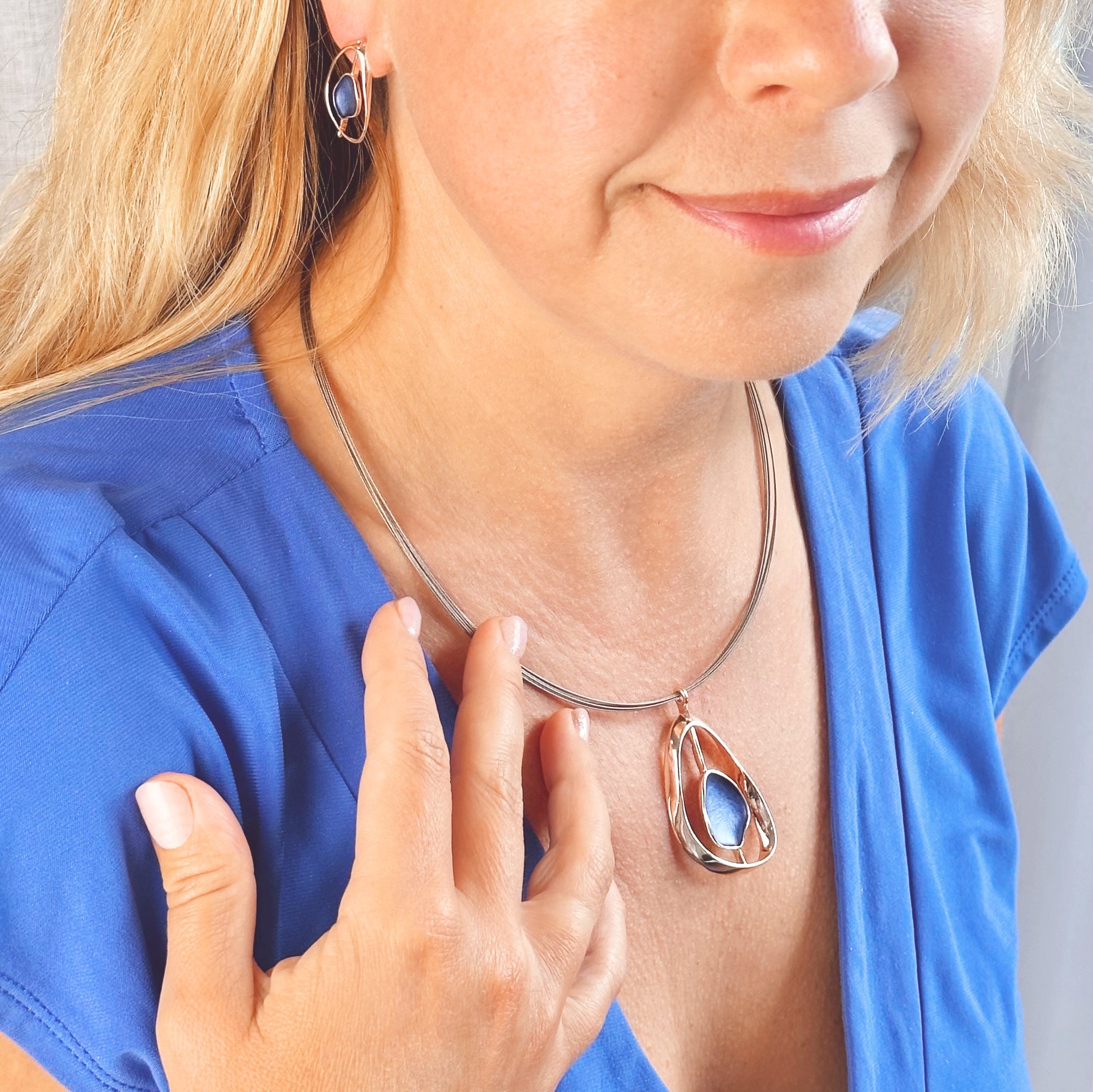 Adjustable Necklace with a brown steel wire chain and an open Pear shape rose gold pendant with a floating textured disc center painted in blue enamel with matching stud earrings worn by a model in a blue blouse