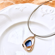 Adjustable Necklace with a brown steel wire chain and an open Pear shape rose gold pendant with a floating textured disc center painted in blue enamel resting on a white ceramic plate on a textured copper background