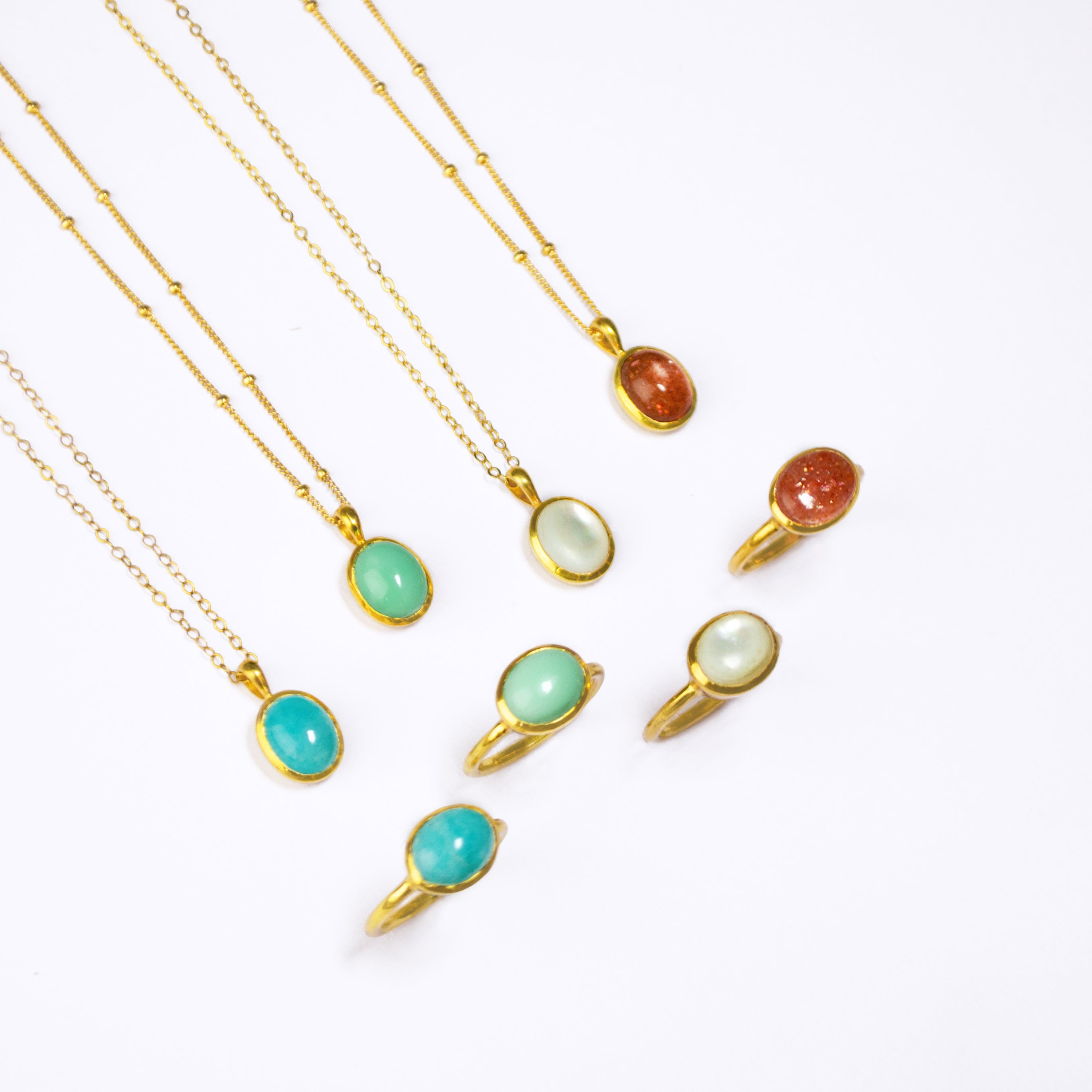 Four Gold Necklaces with oval cabochon pendants with matching Gold rings below. From Left to Right: Blue Amazonite on a cable chain, mint green Chrysoprase on a satellite chain, Mother of Pearl on a cable chain, and Sunstone on a satellite chain