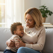 Smiling mom cuddling her laughing toddler on a cozy sofa, wearing a custom birthstone necklace with two colorful teardrop pendants that represent her children’s birth months. Dainty personalized birthstone mom necklace on a simple gold chain, meaningful everyday jewelry and sentimental keepsake, perfect Mother’s Day, birthday, Christmas or baby shower gift idea for moms, new moms and grandmothers who love family-inspired pieces.