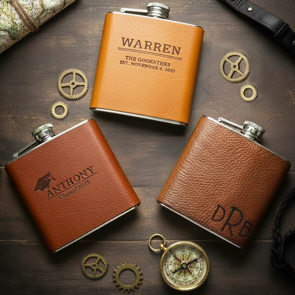 Personalized leather hip flasks in honey brown engraved with names and monograms, styled with compass and vintage props on rustic wood table, custom engraved flask set for groomsmen, best man or father of the groom wedding party gifts
