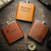 Personalized leather hip flasks in honey brown engraved with names and monograms, styled with compass and vintage props on rustic wood table, custom engraved flask set for groomsmen, best man or father of the groom wedding party gifts