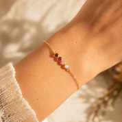 A delicate gold chain bracelet with four small colorful gemstones (red, pink, light pink and white) worn on a person's wrist, set against a warm, softly lit background.