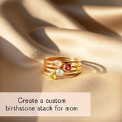 Dainty gold stacking birthstone rings with four colorful gemstones—peridot, aquamarine, garnet and moonstone—arranged in a little stack on shimmering champagne satin, with text “Create a custom birthstone stack for mom,” showcasing personalized family rings for mothers and grandmas.