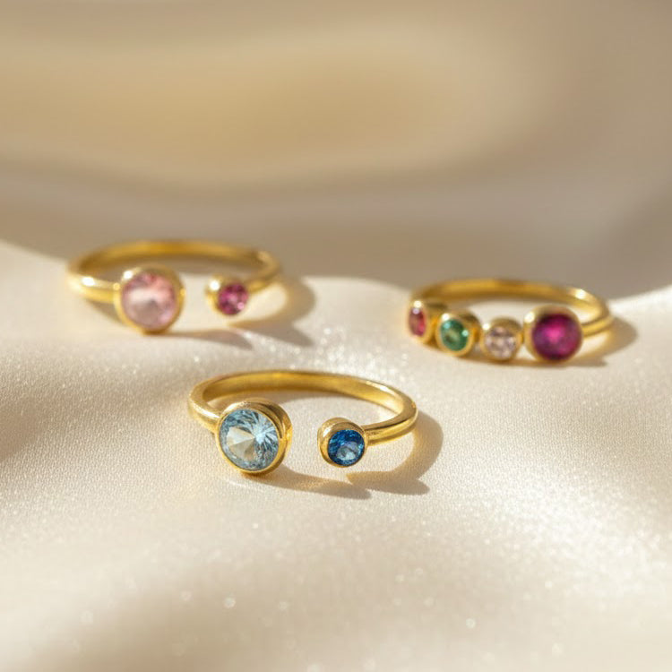Multi birthstone adjustable rings for mom displayed on soft champagne satin, handcrafted in warm gold with bezel-set gemstones in custom family combinations. Front ring features one large aqua blue 5mm birthstone and one smaller blue 3mm stone for a mom of two design, while the others show different child birthstone arrangements. These personalized family rings make meaningful Mother’s Day, birthday or Christmas gifts, celebrating children with colorful, dainty, stackable jewelry.
