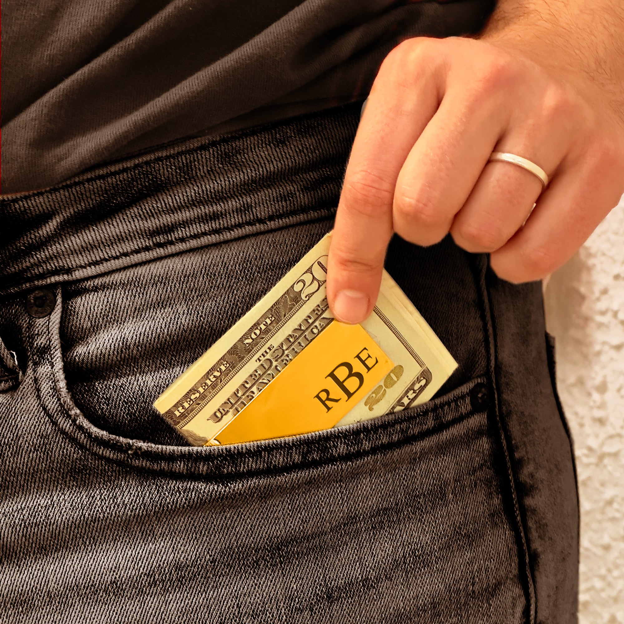 Custom engraved money clip holding folded US twenty dollar bills being slipped into the front pocket of dark denim jeans by a man wearing a simple silver ring, showcasing a sleek personalized metal money clip with initials, a practical minimalist wallet alternative and stylish everyday carry accessory, perfect gift for him, husband, boyfriend, groomsmen or Father’s Day and birthday celebrations.