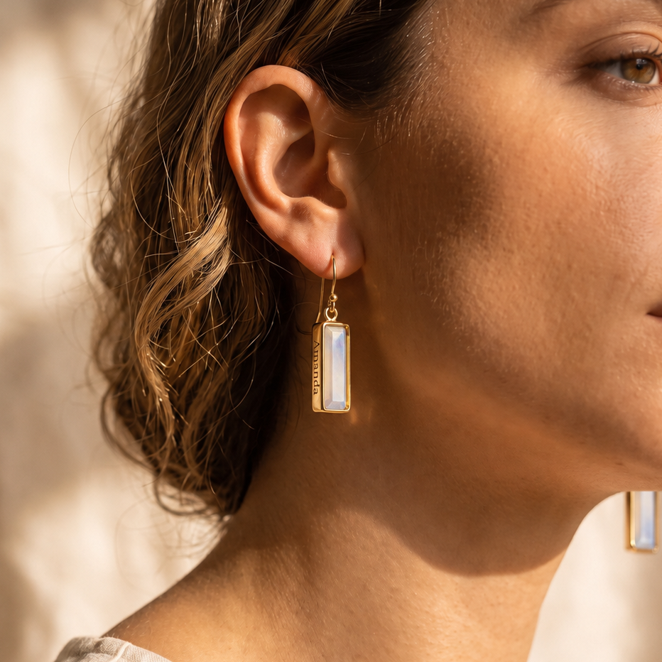 A close-up side profile of a woman wearing a gold geometric drop earring with a rectangular white inlay, set against a warm, sunlit neutral background.