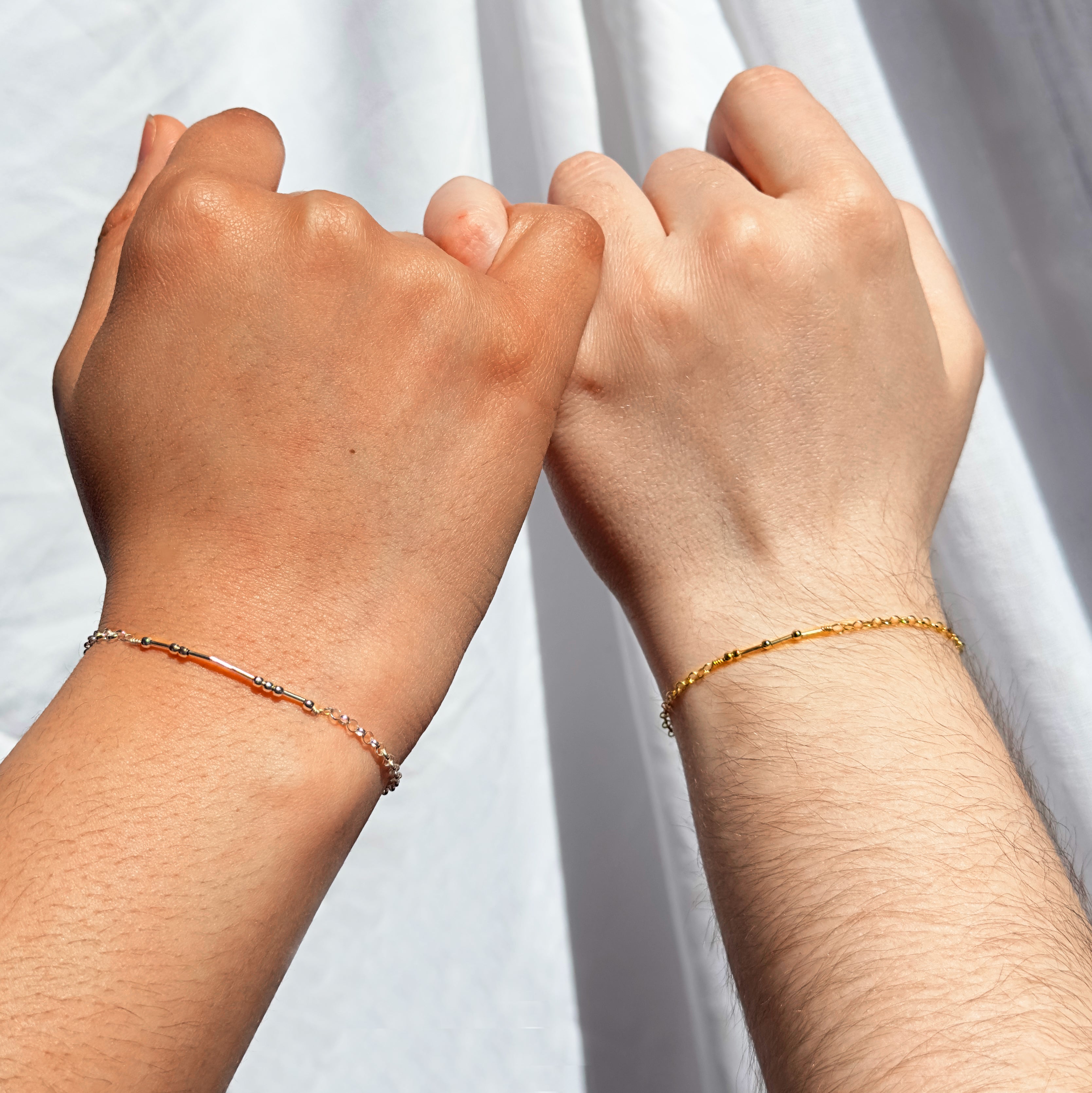 Two people wearing morse code bracelets interlock pinkies against a white fabric background. Personalized couples bracelets, sterling silver code message bracelet, gold filled code bracelet, secret message jewelry, morse code jewelry, jewelry trends for girls, couples jewelry trends