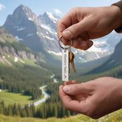 Hands holding a custom engraved bar keychain in front of a scenic mountain valley, with the words “Adventure Awaits” and a small mountain design on the tag, showcasing this personalized travel keychain as a meaningful gift for hikers, campers and outdoor lovers.

