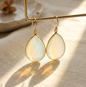 Close-up of opalite teardrop earrings in gold with soft iridescent flashes and warm shadows.