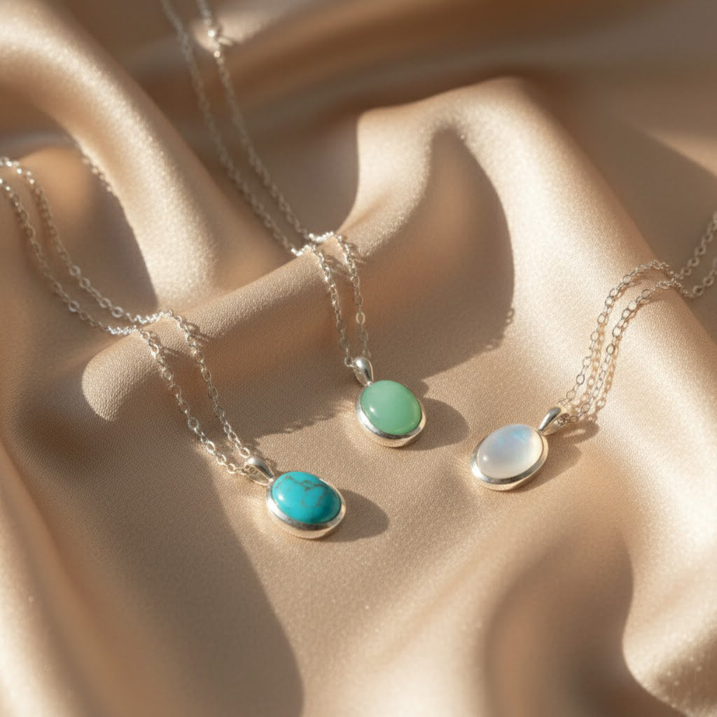 Dainty oval gemstone necklaces in sterling silver displayed on soft champagne satin, featuring three bezel-set stones in turquoise blue, soft green and glowing rainbow moonstone on delicate silver chains. Minimalist gemstone or birthstone pendant necklaces perfect for everyday wear or layering, a meaningful gift for her, mom, sister or best friend for birthdays, Christmas, Mother’s Day, anniversaries or bridesmaid jewelry.