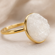 A gold ring featuring a large, rough-cut white gemstone in a bezel setting, photographed on a soft beige textured background.