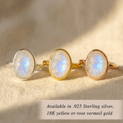 Three oval moonstone rings in silver, yellow gold, and rose gold settings, displayed on a textured beige fabric background with soft shadows. A text overlay at the bottom reads 'Available in .