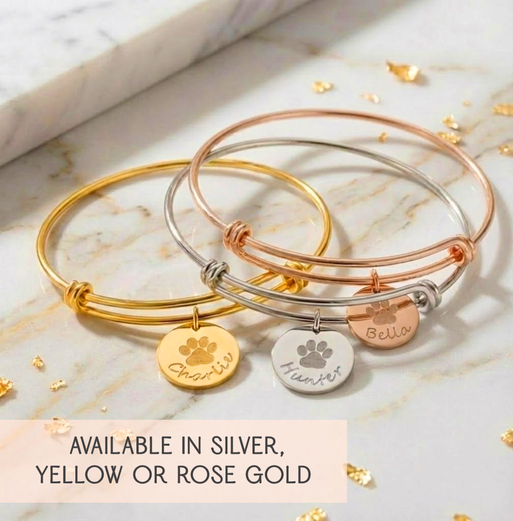 Custom paw print bangle bracelet hanging with soft lights, showcasing engraved pet name and paw print on a polished disc charm. Designed for dog moms, cat moms, and pet lovers who want sentimental personalized jewelry. Adjustable wire bangle fits comfortably for daily wear. Available in sterling silver, yellow gold, or rose gold. Ideal for gifting as pet memorial jewelry, birthday gift, Christmas gift, or meaningful keepsake for women.
