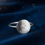 Sterling silver custom paw print ring photographed on a deep midnight blue backdrop with subtle sparkles, emphasizing the engraved paw design and pet name on the polished disc face. Elegant personalized pet memorial ring that can be created from your actual dog or cat paw print, meaningful jewelry for grieving pet parents, dog moms and animal lovers who want a lasting keepsake.