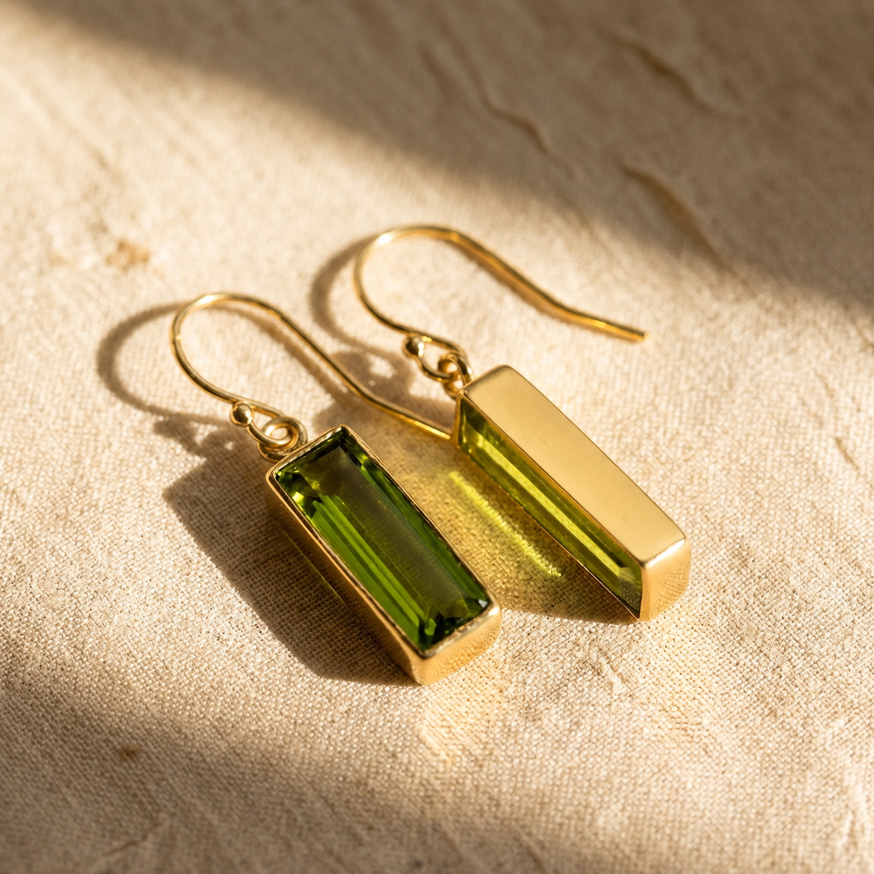 Pair of gold peridot baguette earrings resting on sunlit beige linen