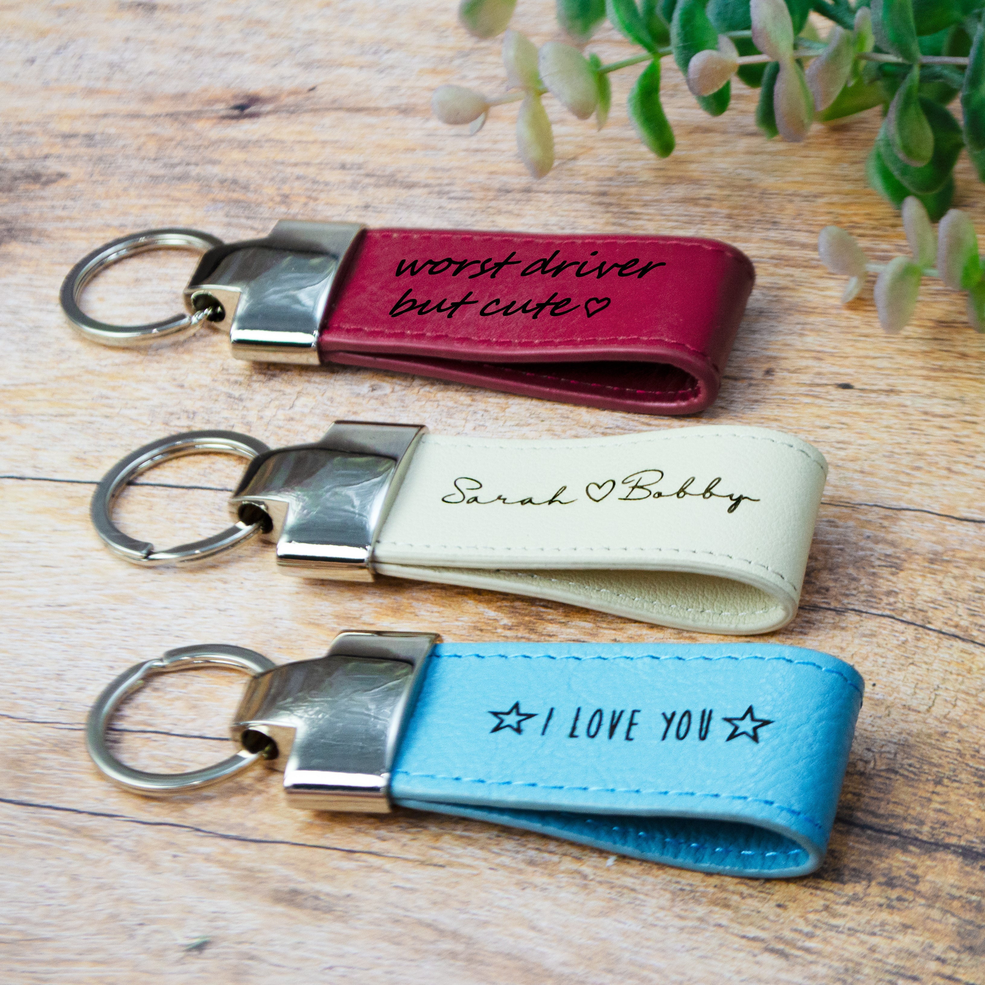 3 natural leather loop keychains in raspberry, white nude, and silver gray blue are all resting against a wood grained background. A green plant can be seen in the upper right corner of the image.  Personalized gift for dad, personalized gift for brother, personalized gift for fiancee, friend, groomsmen, bridesmaids.